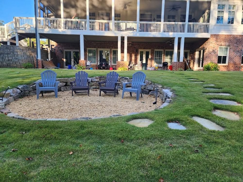 Circular gravel firepit patio with blue Adirondack chairs in a wooded yard